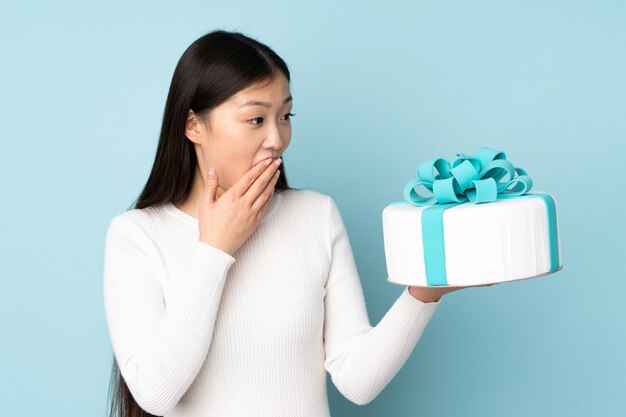 Pastry asian chef holding a big cake isolated on blue space with surprise and shocked facial expression