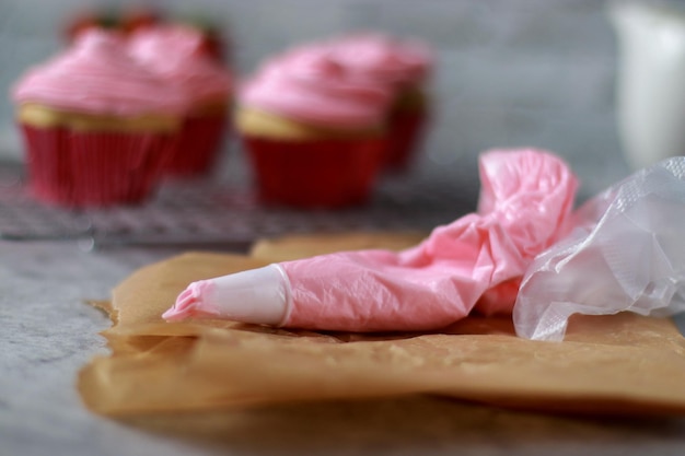 Pastry bag and vanilla muffins with pink strawberry buttercream frosting on top, grey marble table