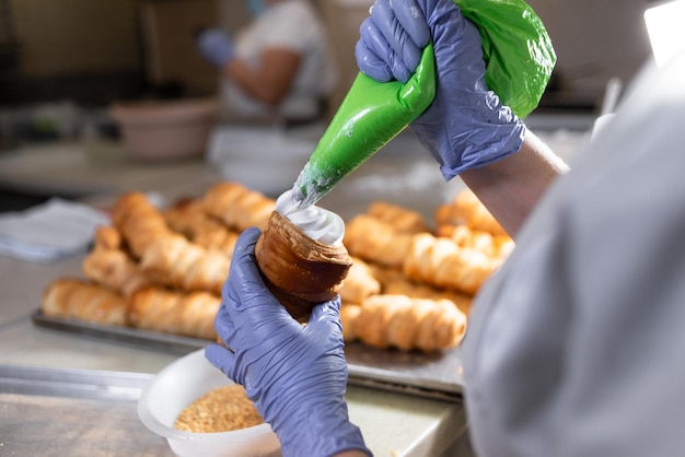 A pastry chef fills a sweet puff pastry tube with whipped proteins from a pastry bag The production process in the kitchen at the bakery Sweet desserts