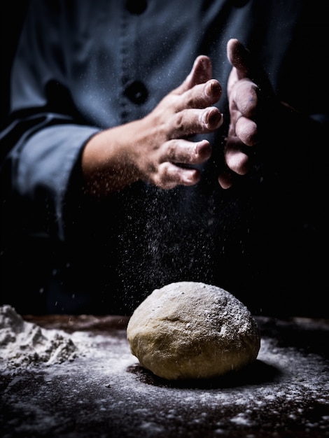 Pastry chef hand sprinkling white flour over Raw Dough on kitchen table.