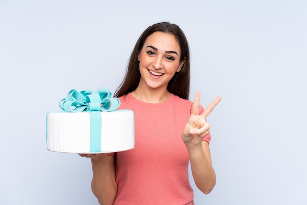 Pastry chef holding a big cake on blue smiling and showing victory sign