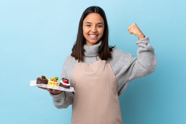 Pastry chef holding a big cake over isolated blue doing strong gesture.