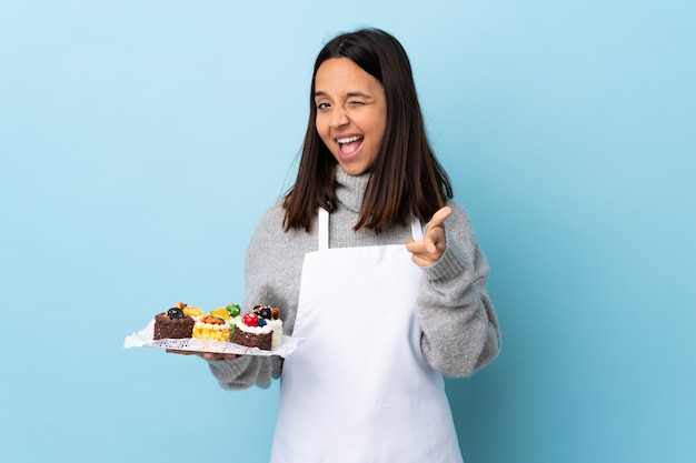 Pastry chef holding a big cake over isolated blue pointing to the front and smiling.