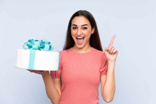 Pastry chef holding a big cake isolated on blue wall pointing up a great idea