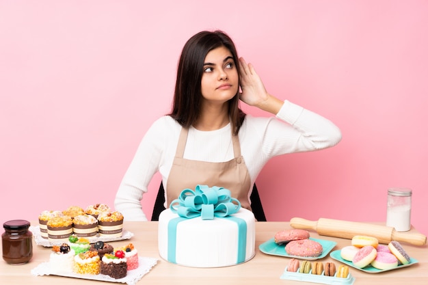 Pastry chef with a big cake in a table over pink wall listening to something by putting hand on the ear