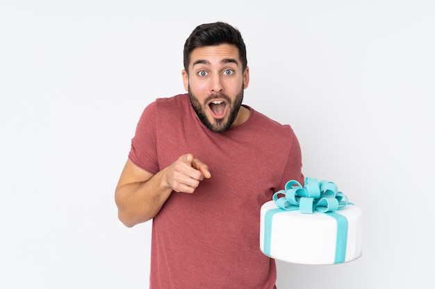 Pastry chef with a big cake on white wall surprised and pointing front