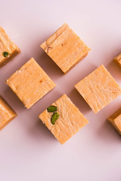 Patisa or Soan Papdi is a popular Indian cube shape flakey and crispy dessert. Served with almonds and pistachio in a plate over moody background. Selective focus