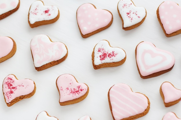 Pattern of gingerbread cookies in the shape of a heart decorated with white and pink glaze on a white wooden surface