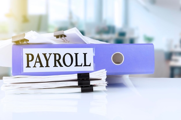 Payroll, the term is written on a folder of documents in trendy purple, lying on a stack of documents on an office table against the backdrop of an office with a soft blurred background. High quality