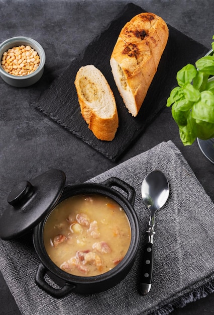 Pea soup with smoked ham in black soucepan on dark background with bread and pepper