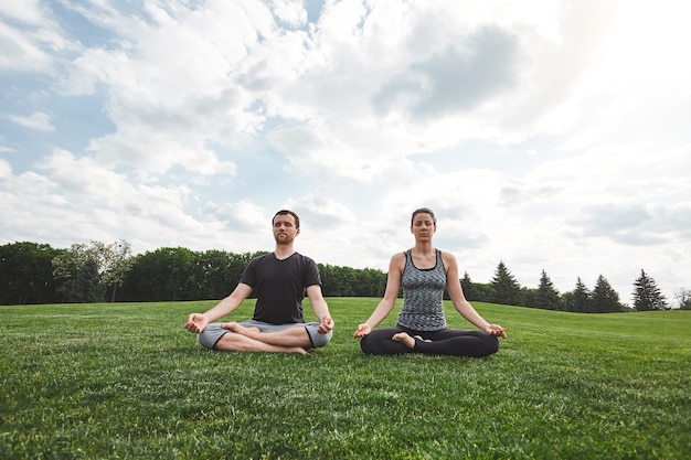 Peace and harmony young couple are practicing yoga outdoors while sitting on a green lawn