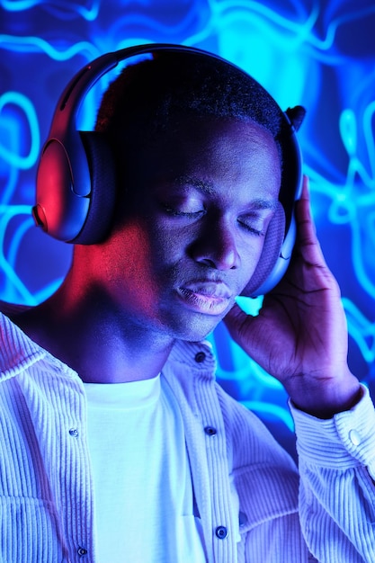 Peaceful African American male wearing casual clothes listening to playlist in wireless headset against blue neon illumination