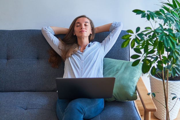 Peaceful calm woman with hands behind head resting alone on comfortable sofa with computer on knees at cozy comfy living room at home among indoors plants