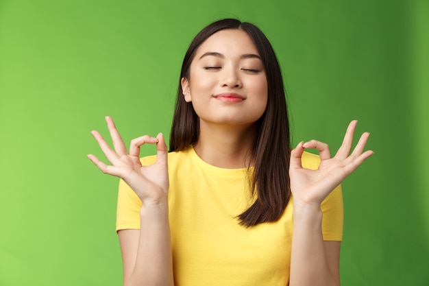 Peaceful charming asian girl brunette inhale fresh air, relaxing breathing happily, close eyes smiling, hold hands zen mudra signs reach nirvana, meditation, practice yoga, green background.