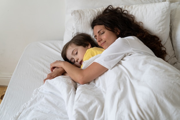 Peaceful loving mum hug preschool son sleeping together with child in cozy bed under warm blanket