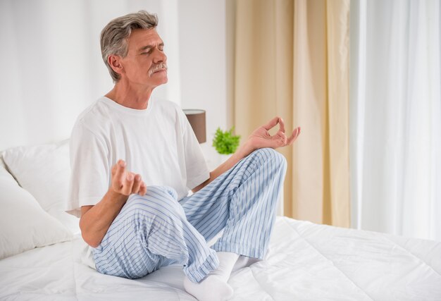 Peaceful senior man meditating seated on a bed.