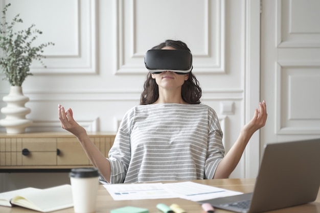 Peaceful woman employee meditating in glasses of virtual reality sitting in zen pose at workplace