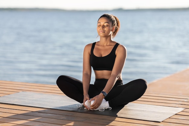 Peaceful young black woman meditating next to city lake