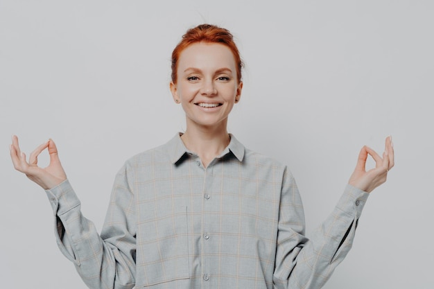 Peaceful young smiling redhead woman holding hands in mudra isolated on grey studio background