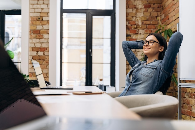 Peaceful young woman feeling relaxed at work desk with closed eyes and holding hands behind head in office interior