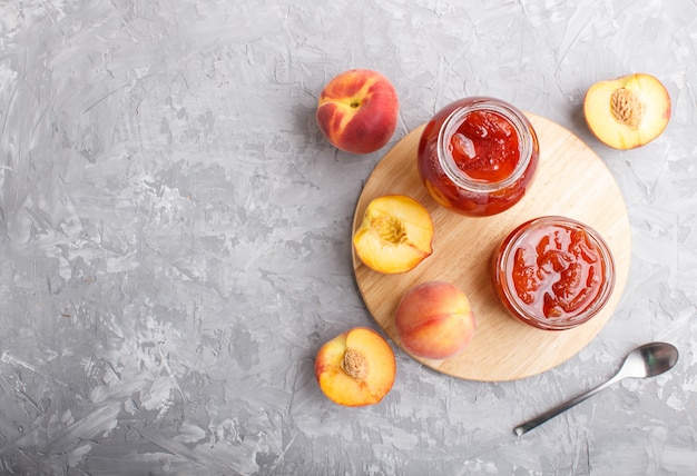 Peach jam in a glass jar with fresh fruits on gray concrete background