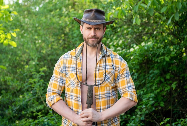 Peasant holding pitchfork outdoors Unshaven guy wearing peasant hat and plaid shirt Peasant man