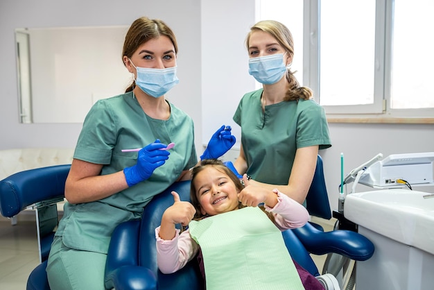 Pediatric female dentist with assistant with child girl patient showing thumbs up