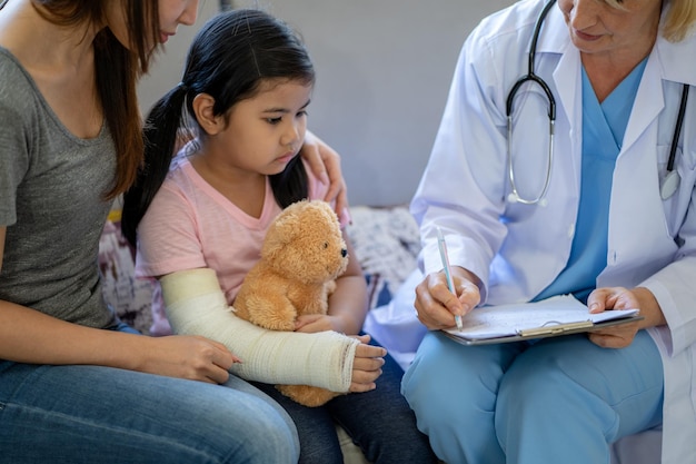 Pediatrician doctor examining little asian girl with a broken arm wearing a cast at hospital