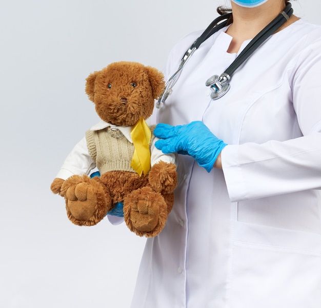 Pediatrician in white coat, blue latex gloves holds a brown teddy bear with a yellow ribbon on a sweater