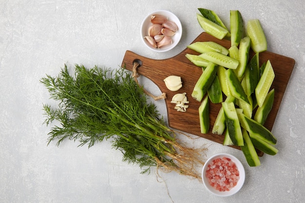 Peeled fresh cucumbers on a brown wooden cutting board with a bunch of dill garlic and pink salt on a light gray concrete table