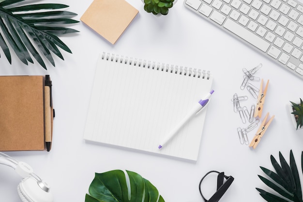 Photo pen and blank spiral notepad surrounded with office stationeries on white desk