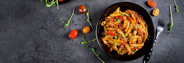 Penne pasta in tomato sauce with meat, tomatoes decorated with pea sprouts on a dark table.