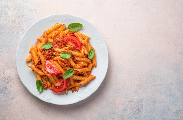 Penne pasta with bolognese sauce, fresh tomatoes and basil in a white plate on a light pink background. Top view, copy space.