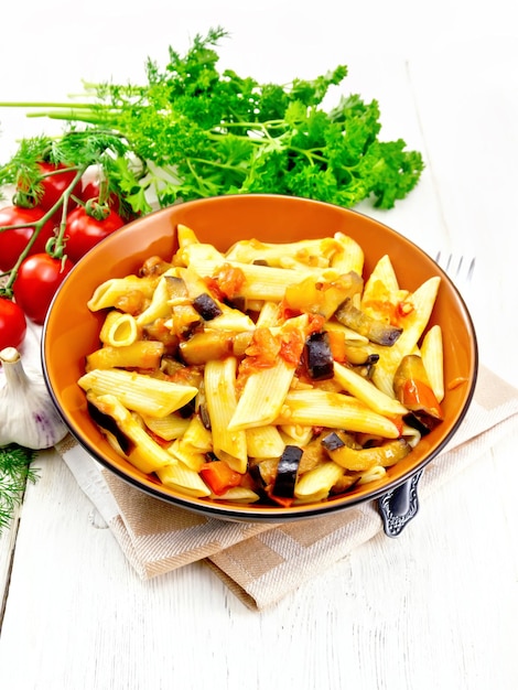 Penne pasta with eggplant and tomatoes in a bowl on napkin, fork, garlic and parsley on light wooden board background