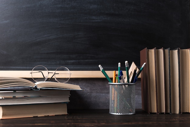 Pens, pencils, books and glasses on the table, against the background of a chalkboard