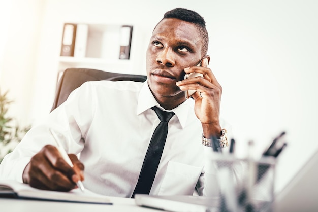 Pensive African businessman is talking on smartphone and writing in notebook in modern office.