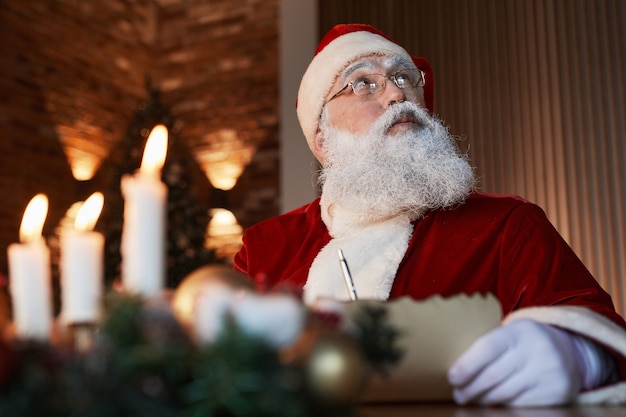 Pensive aged santa claus in eyeglasses sitting at table and looking up while writing letter