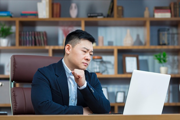 Pensive and anxious businessman in a classic office works at a desk with a laptop an Asian makes important decisions chooses a business strategy