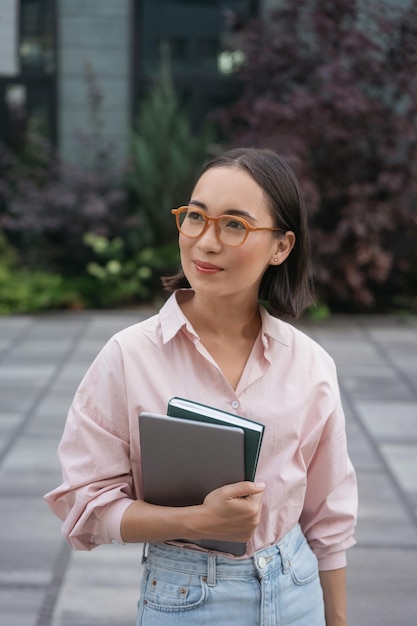 Pensive asian student wearing stylish eyeglasses holding book, laptop standing in university campus