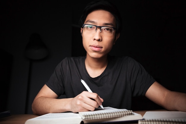 Pensive asian young man in glasses studying and writing at the table in dark room