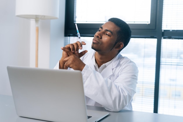 Pensive black male doctor in white uniform thoughtful looking to syringe with vaccine in hand sitting at table with laptop in clinic on background of window