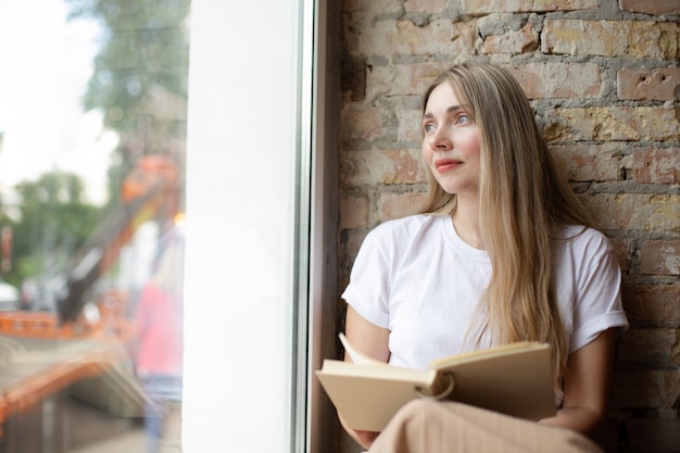 Pensive blonde caucasian woman in a white t-shirt looks out the large window reading a book leaning on a brick wall