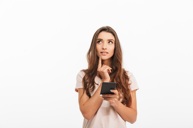 Pensive brunette woman in t-shirt holding smartphone and looking up over white wall