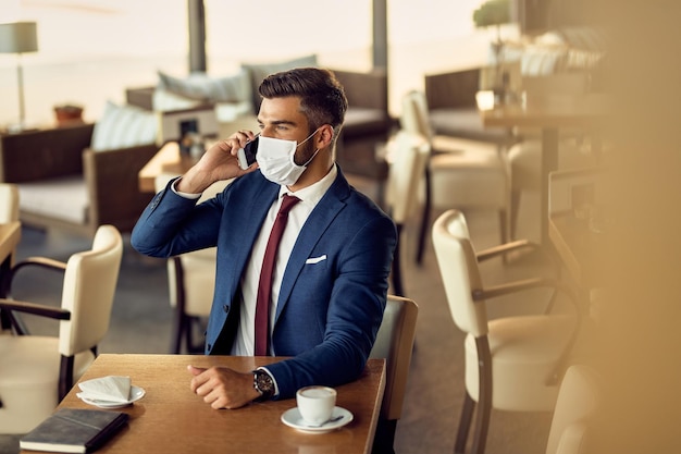 Pensive businessman talking on the phone and wearing protective face mask in a cafe