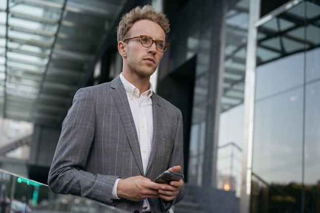 Pensive businessman wearing suit stylish eyeglasses holding mobile phone walking on the street