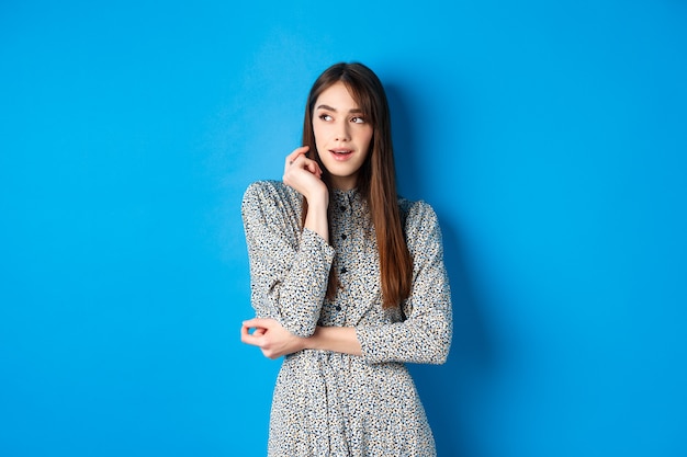 Pensive caucasian girl with long natural hair, wearing vintage dress, looking left with thoughtful face, standing on blue.