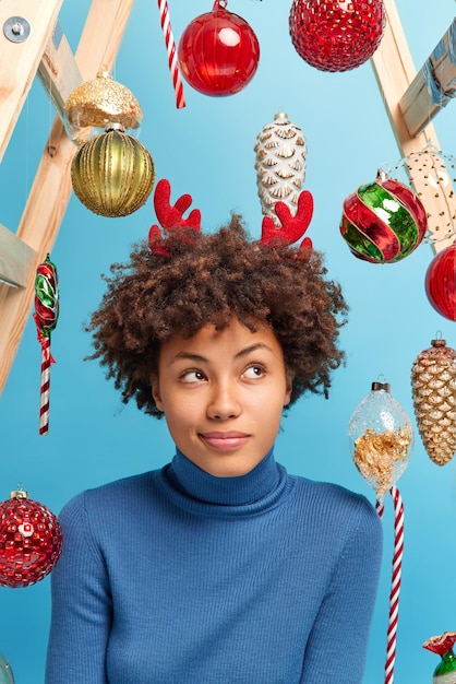 Pensive curly haired woman dressed casually looks away thoughtfully considers something in mind poses around new year toys isolated over blue background thinks about winter holidays traditions