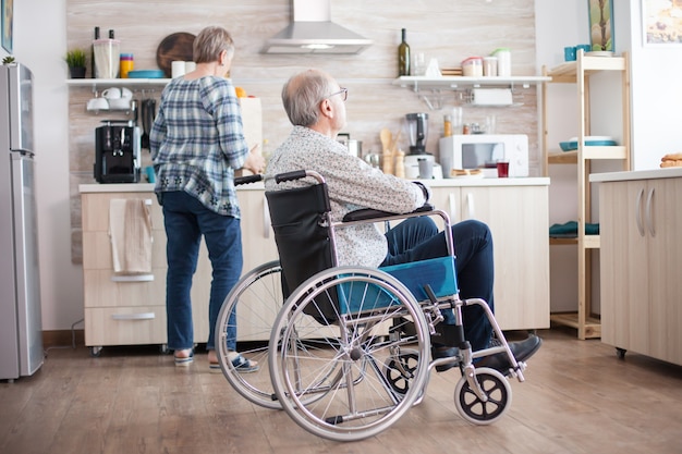 Pensive disabled man in wheelchair looking through kitchen window while wife is unpacking food from grocery bag. Pensioner, parallysis, invalid, handicapped.