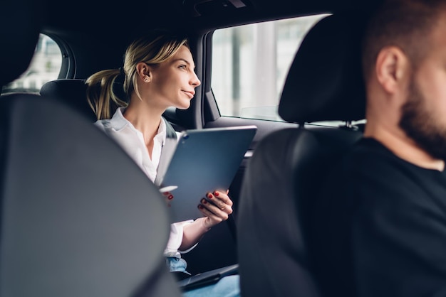 Pensive female passenger with portable tablet for remote working sitting at automobile backseat