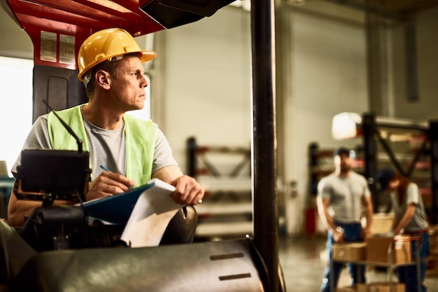 Pensive forklift operator writing on clipboard while working in industrial warehouse